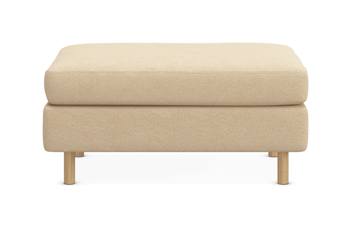 Beige puf/ottoman med stofbetræk og lyse træben, fotograferet direkte forfra på hvid baggrund.