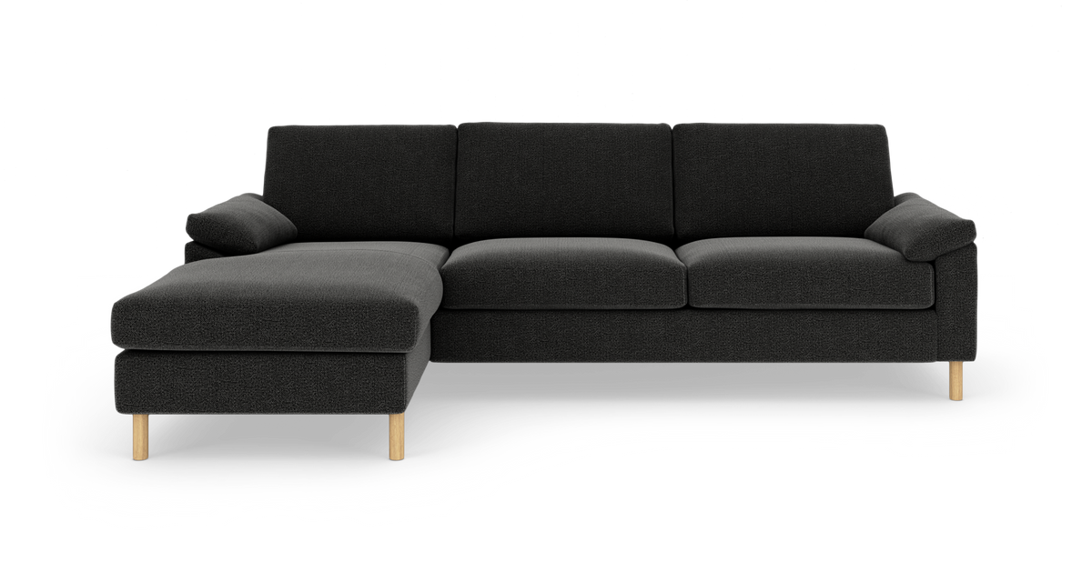 Sort hjørnesofa med chaiselong til venstre og lyse træben, fotograferet lige forfra på hvid studiobaggrund.
