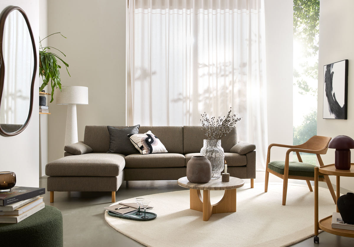 Lyst stue med beige, lette gardiner, gråbrun hjørnesofa, rundt sofabord i træ/sten med vaser og et lys, grøn trælænestol, spejl og gulvlampe i neutrale nuancer.
