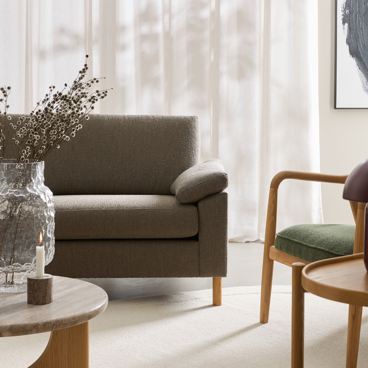 Neutralt stuemiljø med taupefarvet sofa, trælænestol med grønt sæde, rundt sofabord i træ/sten, riflet glasvase med grene og et tændt stearinlys foran lyse gardiner.