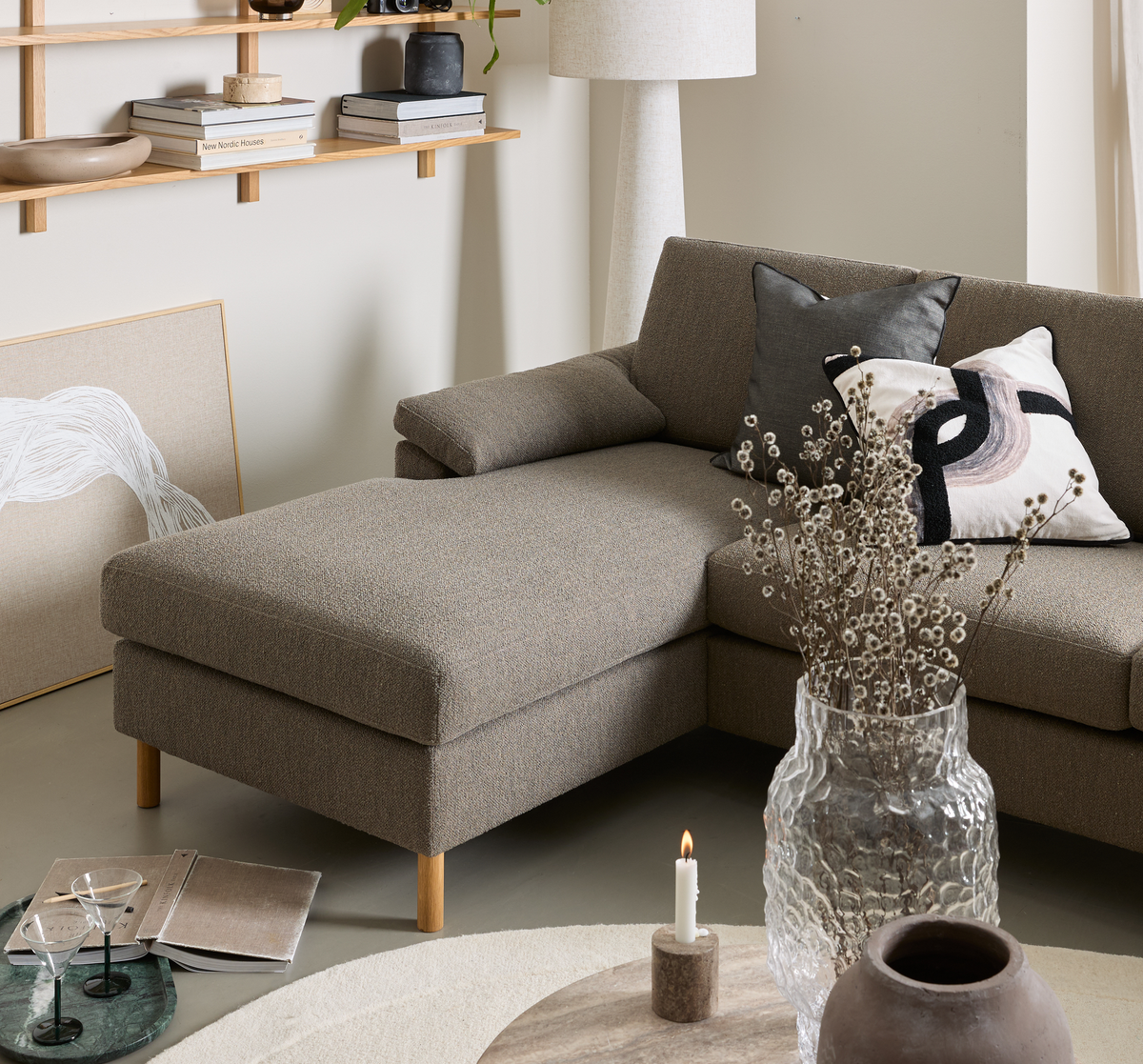 Beigegrå hjørnesofa med puder, glasvase med tørrede grene på rundt sofabord og tændt lys; gulvlampe og væghylde med keramik og bøger, bl.a. New Nordic Houses, i en rolig skandinavisk stue.