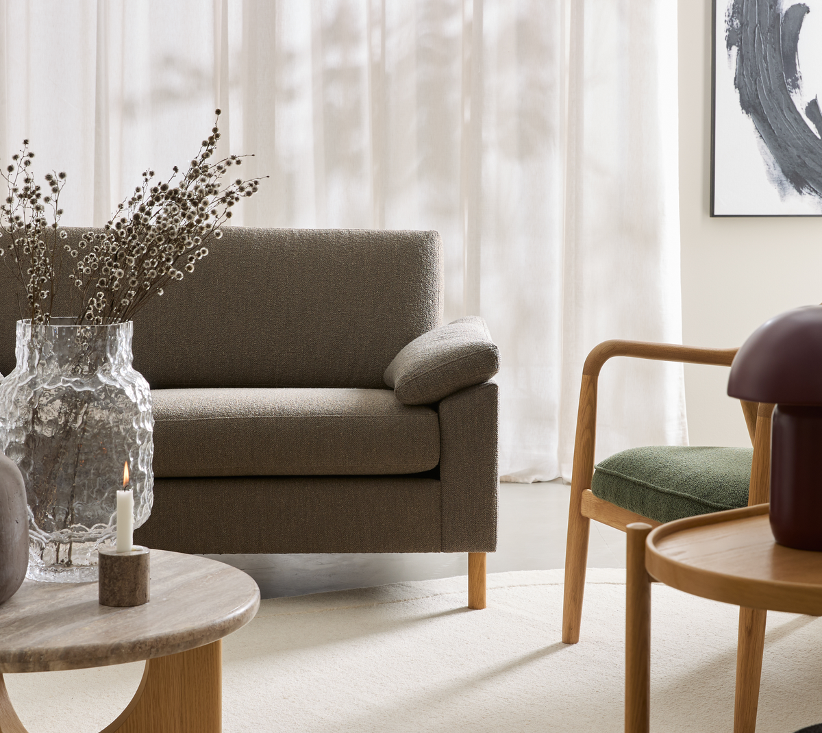 Lyst, minimalistisk stue: beige sofa, trælænestol med grøn sædehynde, runde sofaborde, tændt lys og tekstureret glasvase med tørrede grene foran lette gardiner og abstrakt vægkunst.