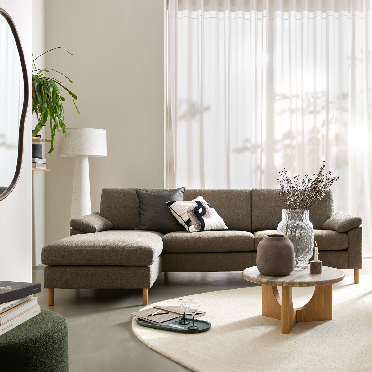 Lys stue med taupe hjørnesofa, rundt sofabord i træ/sten, vaser og lys, gulvlampe, plante og lette gardiner i dagslys.