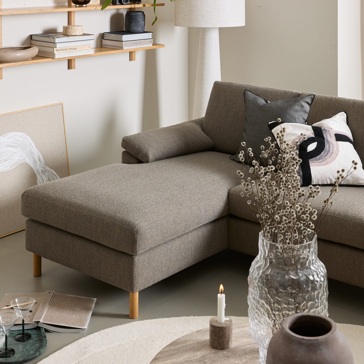 Beigegrå hjørnesofa med træben, grafiske puder, glasvase med tørrede grene og tændt lys på sofabord; på væggen en hylde med bøger som New Nordic Houses og en lys gulvlampe i en lys stue.