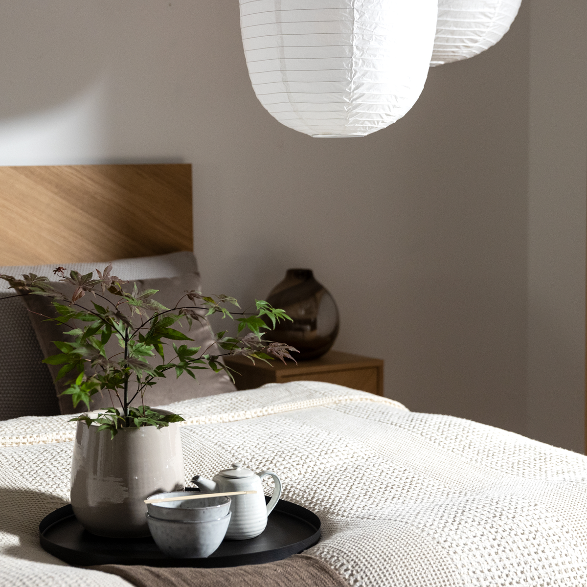 Soveværelse med trægavl, hvide rislamper og en bakke på sengen med tekande, skåle og en potteplante på en strikket beige plaid.