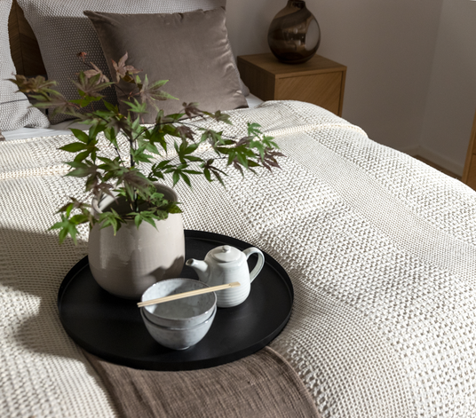 Redt seng i neutrale toner med en sort bakke med beige vase med grønne kviste, en lille hvid tekande og en skål.