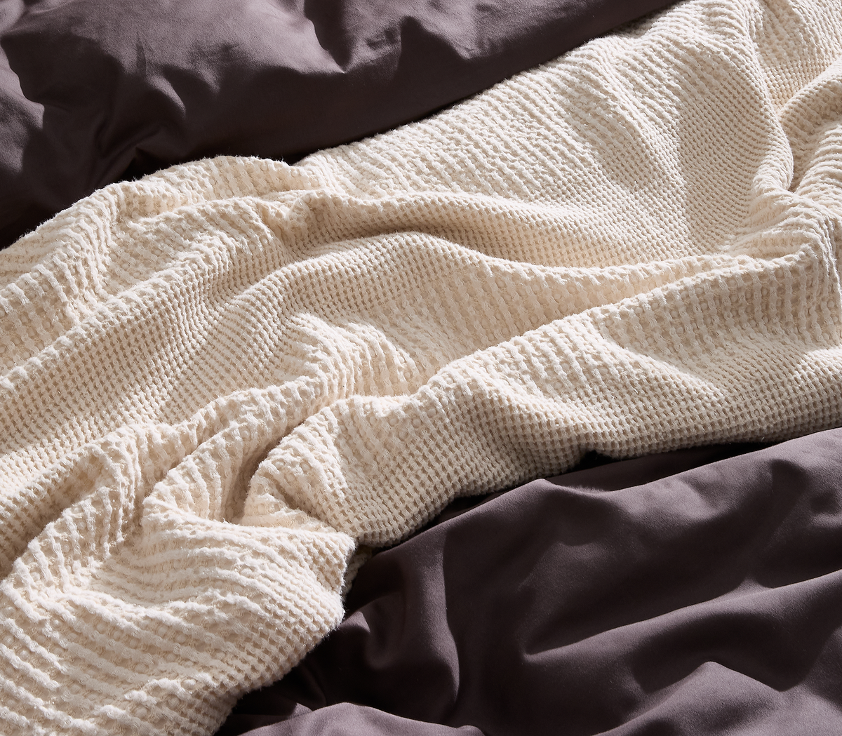 Beige vaffelstrikket plaid på mørkegråt sengetøj i blødt dagslys.