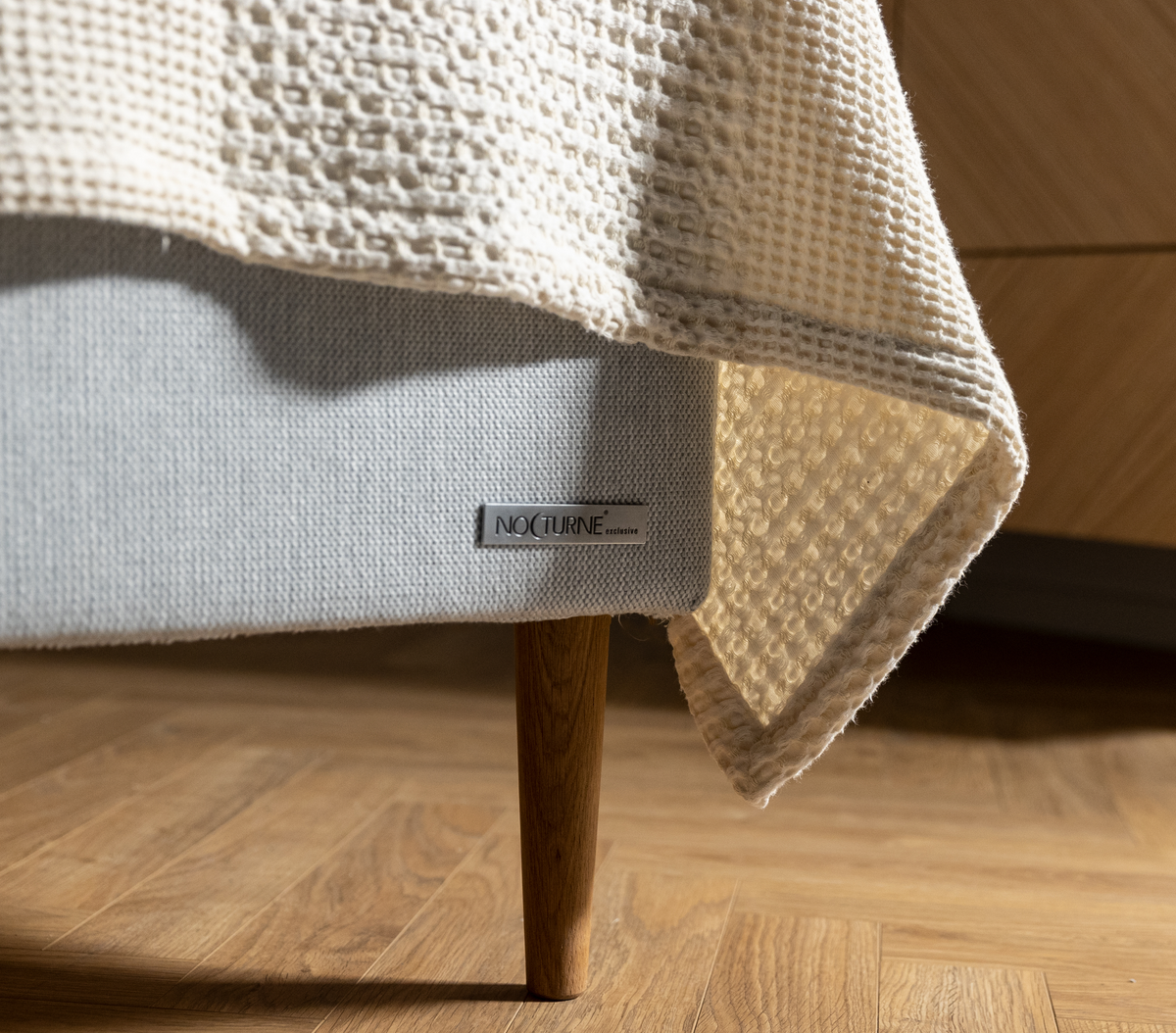 Nærbillede af en lysegrå sengebund med træben og en beige vaffelstrikket plaid over kanten, metalplade NOCTURNE exclusive.