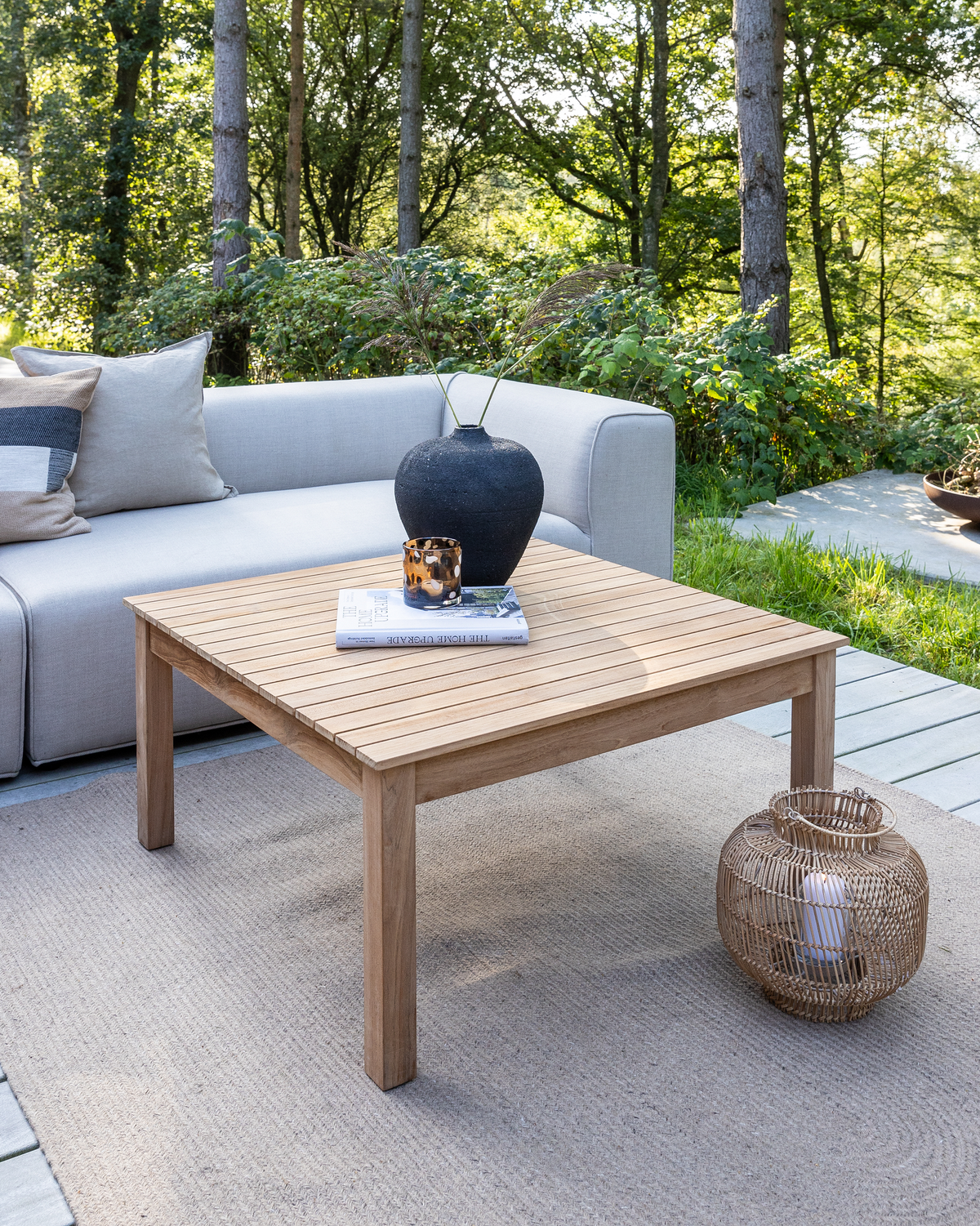 Udendørs lounge med teak sofabord på tæppe, grå sofa, sort vase med græs, bog og lille lysglas på bordet samt en rattanlanterne med lys ved siden af, omgivet af grønne træer.