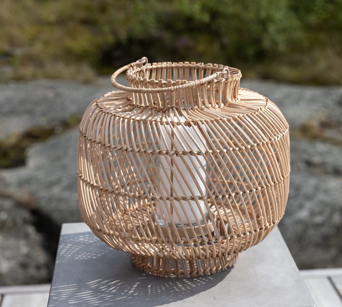 Rund lanterne i rattan med glasindsats og hvidt lys, placeret udendørs på et bord.