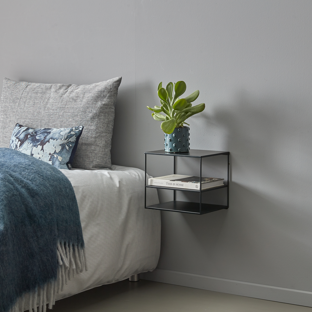 Soveværelse med vægmonteret sort sengebord med en krukkeplante og bogen ”THIS IS HOME”, ved siden af en grå seng med blå plaid og mønstret pude.