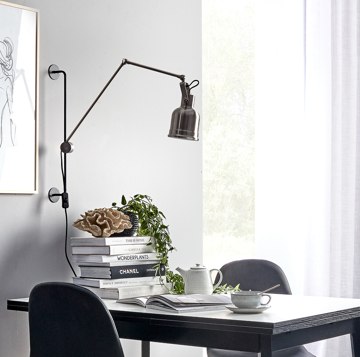 Skandinavisk spise- eller arbejdsplads: sort bord ved lyst vindue med vægmonteret svingarm-lampe over en stak bøger (This Is Home, Wonderplants, Chanel), tekande og kop, grøn plante og stregtegning på væggen.