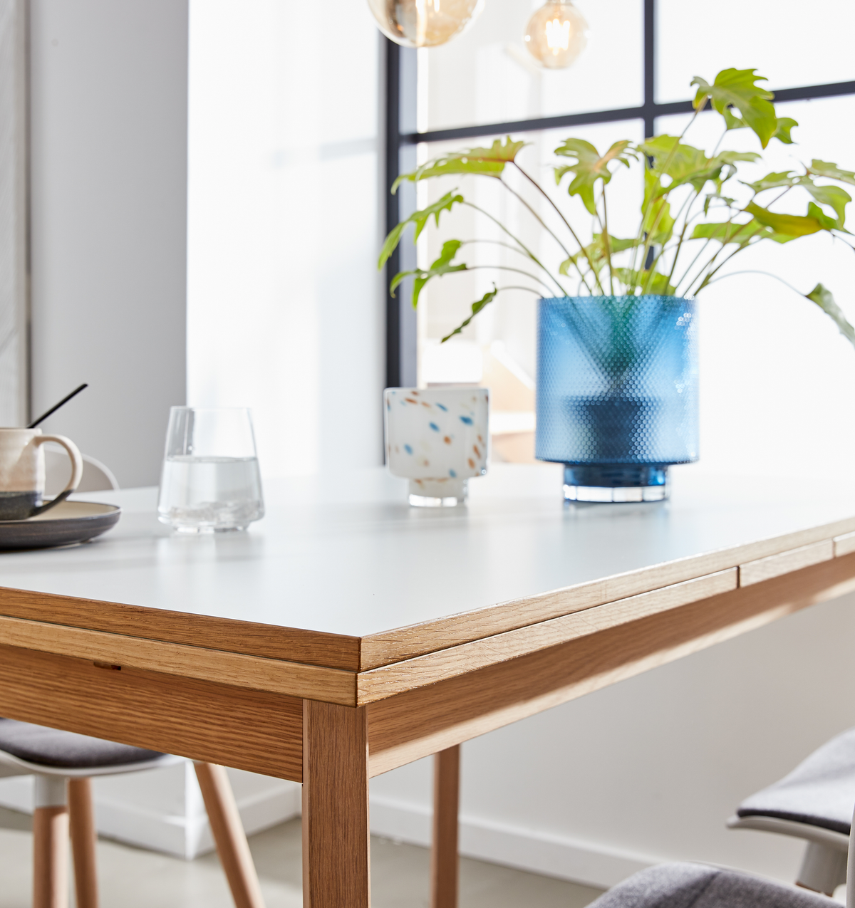 Lyst spisebord med hvid bordplade og egetræskant, blå glasvase med grøn plante, vandglas og kop i et solrigt rum med sorte vinduesrammer.