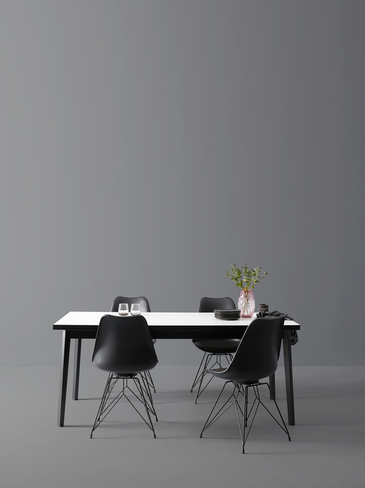Minimalistisk spisebordsscene med hvid bordplade og fire sorte stole; på bordet er to glas, en stak mørke tallerkener og en vase med grønne grene.