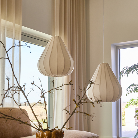 To beige pendellamper i stof i en lys skandinavisk stue med bløde gardiner, vindueslys og en gren i vase.