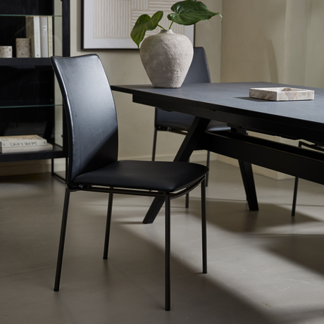 Sort spisebordsstol ved mørkt bord, med vase og grøn plante på bordet i et moderne, lyst rum.