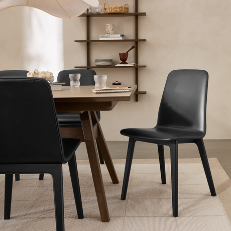 Moderne spiseområde med sorte stole omkring et mørkt træbord, beige væg og åbne hylder med pynt, blødt dagslys.