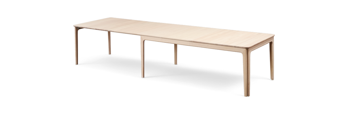 Lyst og minimalistisk hjemmekontor med træskrivebord, computerskærm, tastatur, sort bordlampe, planter og hylder med dekorationer mod en hvid væg.