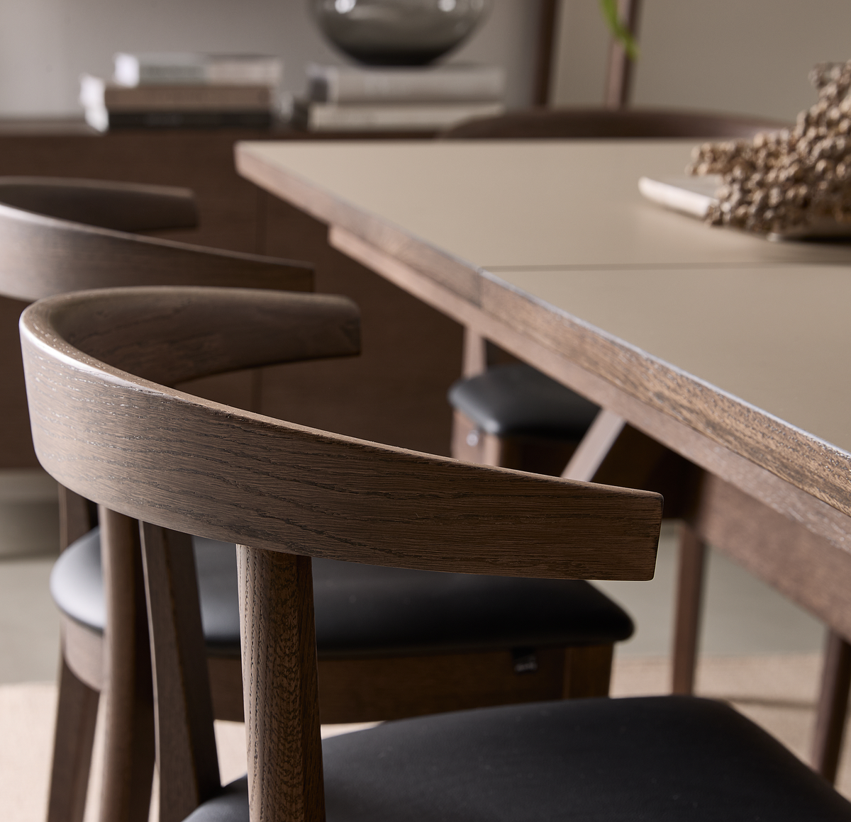 Nærbillede af en moderne spisebordsstol i mørkt træ med buet ryglæn og sort sæde ved siden af et spisebord i træ i et lyst, minimalistisk spiseområde.