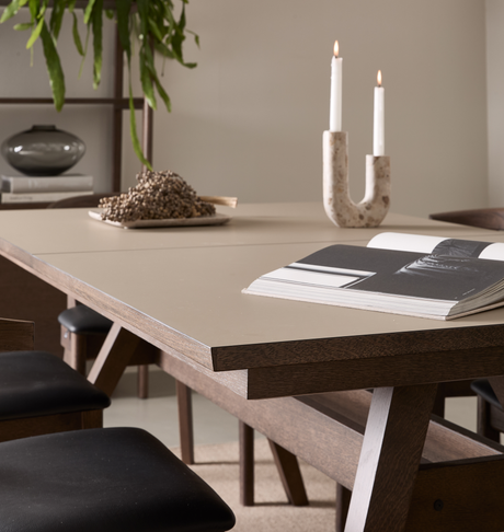 Spisebord i træ med beige bordplade, åben bog og tændt U‑formet lysestage; hylde med vase og hængende plante i baggrunden.