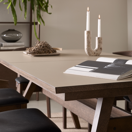 Nærbillede af et moderne spisebord i træ med beige bordplade, et åbent magasin, en U-formet lysestage med to tændte lys og et dekorativt fad i et roligt hjemmemiljø.