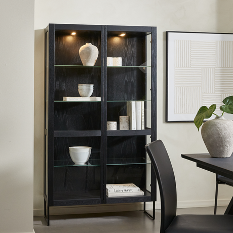 Sort vitrineskab med glashylder og indbygget lys, stylet med keramik, bøger (Chopin Wagner) og en grøn plante; ved siden af spisebord og vægkunst i en lys, moderne indretning.