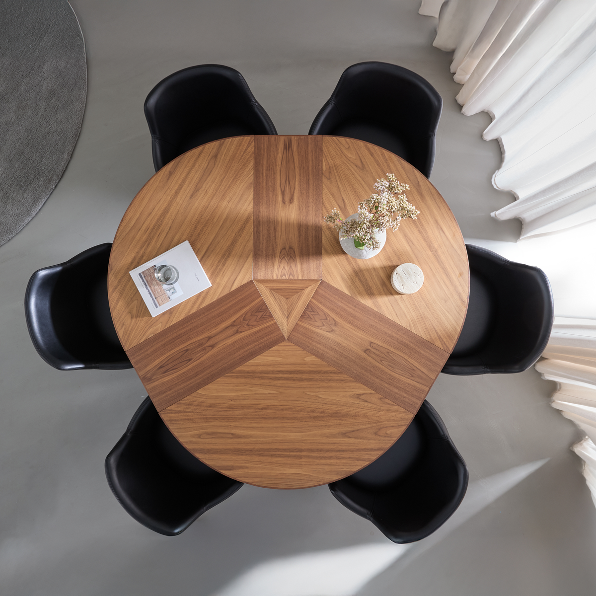 Rundt spisebord i valnød med geometrisk intarsia set ovenfra, omgivet af fem sorte stole, med en vase med blomster, en lille stenunderlag og et magasin ved lyse gardiner.