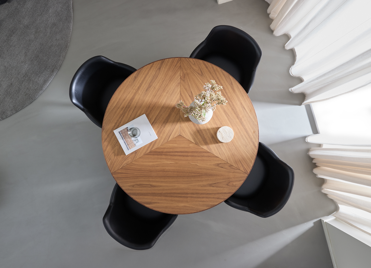 Rundt spisebord i træ set ovenfra med tre sorte stole, en vase med lyse blomster, en lille rund coaster og et magasin i blødt dagslys ved hvide gardiner.