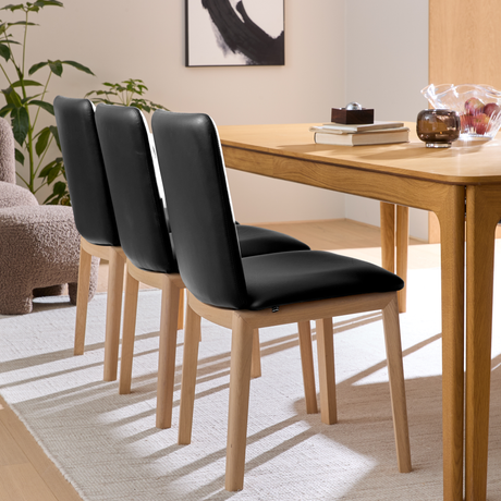 Tre sorte polstrede spisebordsstole med lyse træben ved et egetræsbord på et lyst tæppe i en lys, skandinavisk spisestue.