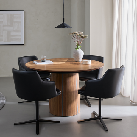 Rundt spisebord i træ med sorte stole, pendellampe og vase med tørrede blomster i et lyst, minimalistisk spiseområde.