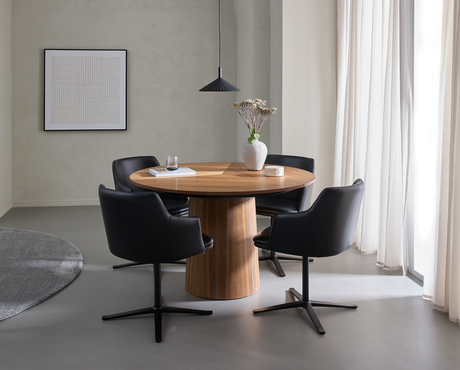 Lyst spisestue med rundt valnødsbord, fire sorte drejestole, en sort pendel og en vase med tørrede blomster; bløde hvide gardiner ved vinduet og minimalistisk vægkunst.