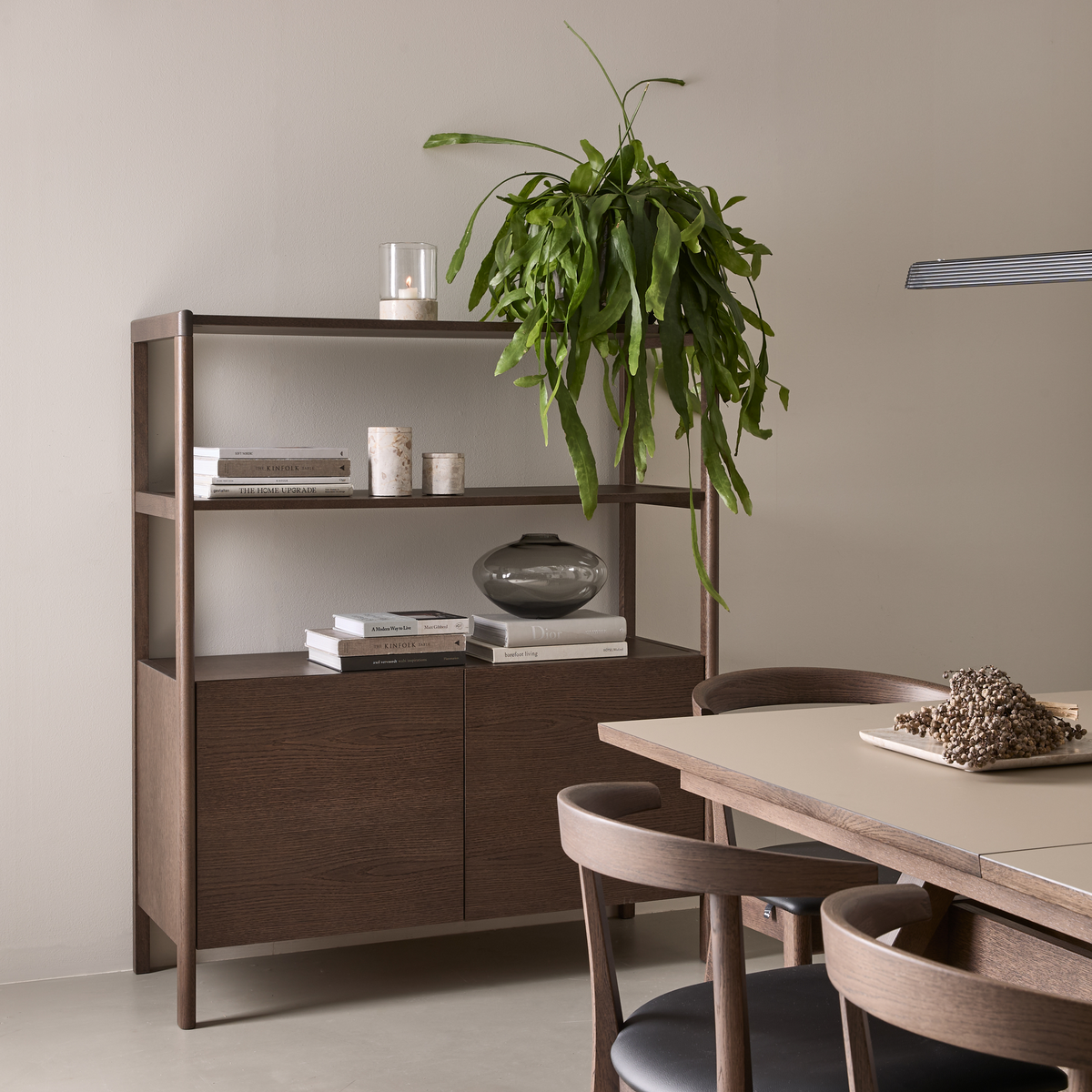 Roligt spiseområde i skandinavisk stil med mørkt træskab og åbne hylder stylet med bøger, lys, glasvase og hængende grøn plante ved siden af bord og stole; en bog har titlen Dior.