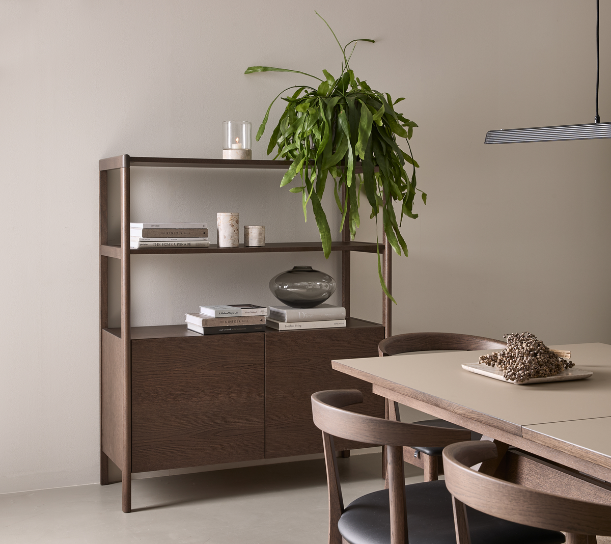 Skandinavisk spisestue med mørkbejdset træbord og stole, åben reol med bøger, lys og vase, hængende grøn plante og slank pendel i blødt dagslys.