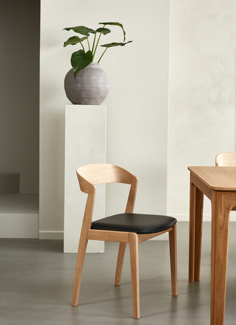 Træstol med sort sæde ved siden af et spisebord; rund vase med grøn plante på sokkel i en lys, minimalistisk spisestue.