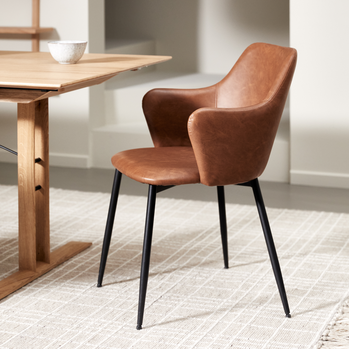 Brun læderbetrukket spisebordsstol med sorte metalben ved siden af et lyst træbord på et beige, tekstureret tæppe i et lyst, minimalistisk rum.