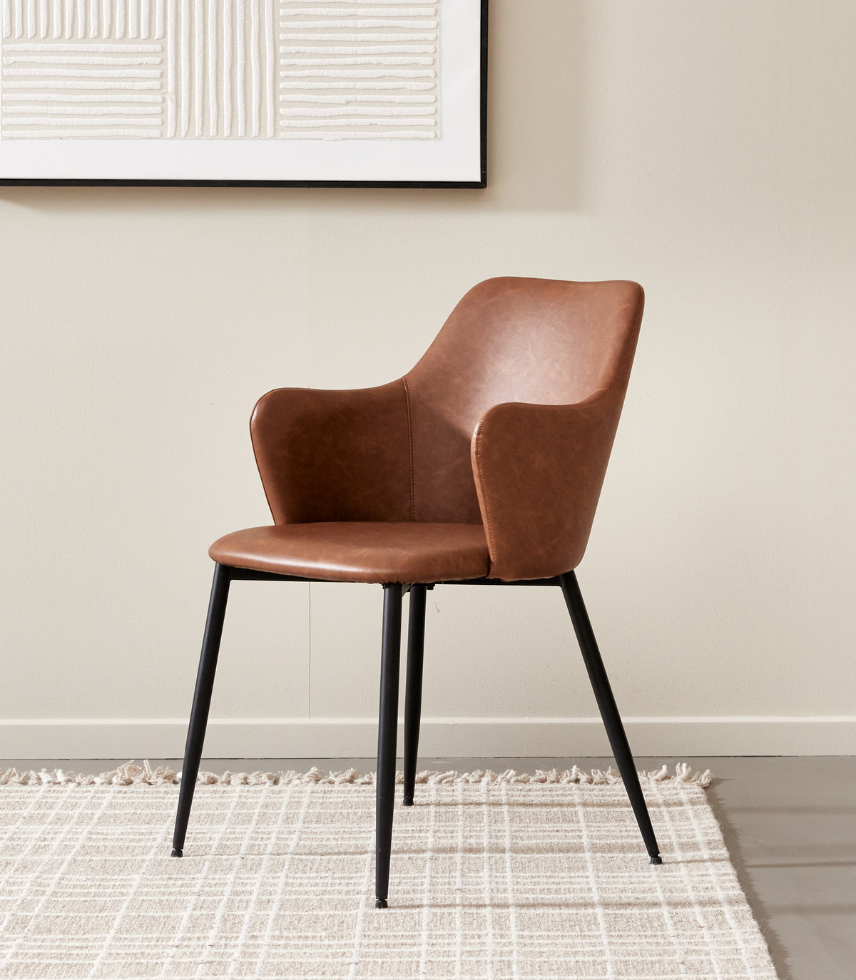Brun læderlook stol med armlæn og sorte metalben på lyst ternet tæppe mod beige væg med minimalistisk vægkunst.