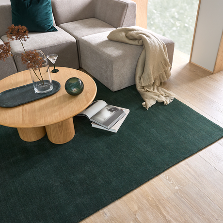 Lyst hjørne af stue med grøn tæppe, rundt sofabord i lyst træ med vase og dekoration, beige modulsofa med plaid og en opslået bog på tæppet.