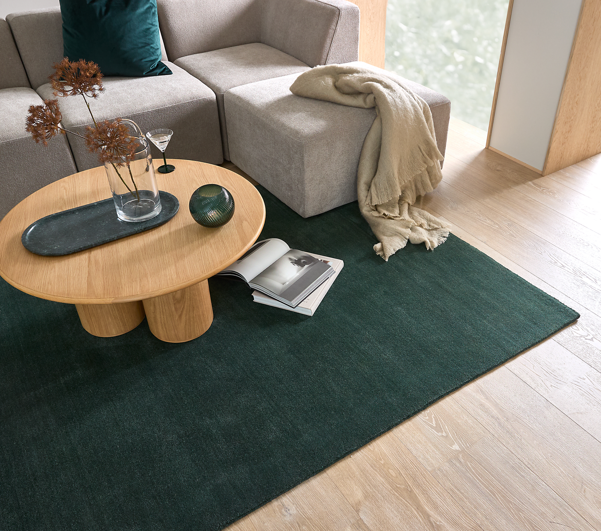 Moderne stue med beige modulsofa, rundt sofabord i eg og mørkegrønt tæppe; et opslået magasin, glasvase med tørrede grene og et plaid ved vinduet.