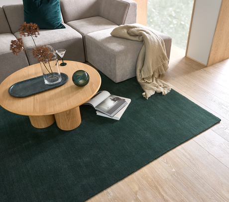 Moderne stue med beige modulsofa, rundt sofabord i eg og mørkegrønt tæppe; et opslået magasin, glasvase med tørrede grene og et plaid ved vinduet.