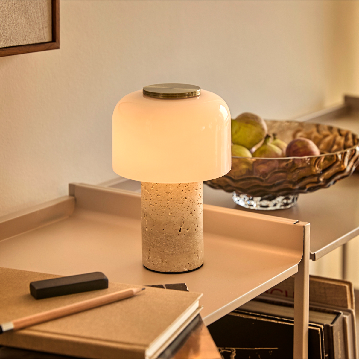 Bordlampe med lys glas-skærm og cylindrisk stenfod på beige hylde, frugtskål i baggrunden.
