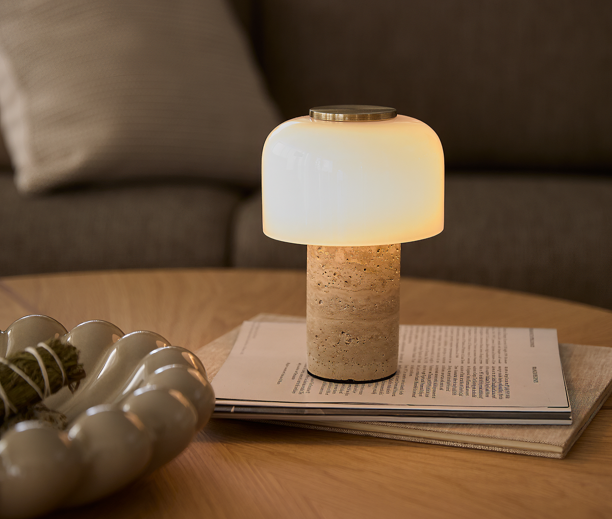 Varmt lysende bordlampe med hvid glaskuppel og cylindrisk stenfod på en stak magasiner på et træbord, med sofa og dekorationsskål i baggrunden.