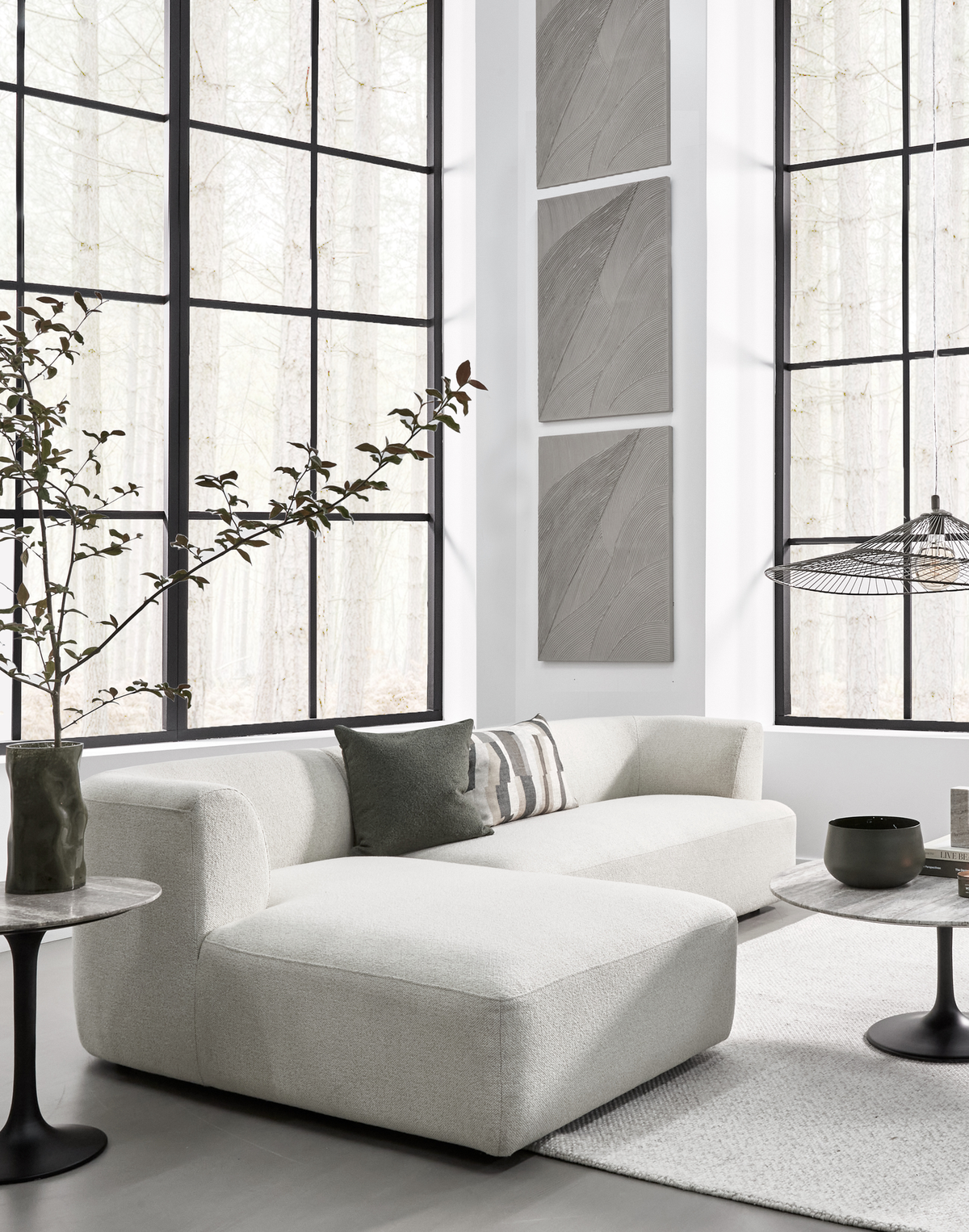 Lys, minimalistisk stue med lys hjørnesofa, store sorte sprossede vinduer, neutralt tæppe, runde piedestalborde, grønne detaljer og tre smalle vægpaneler med kunst.