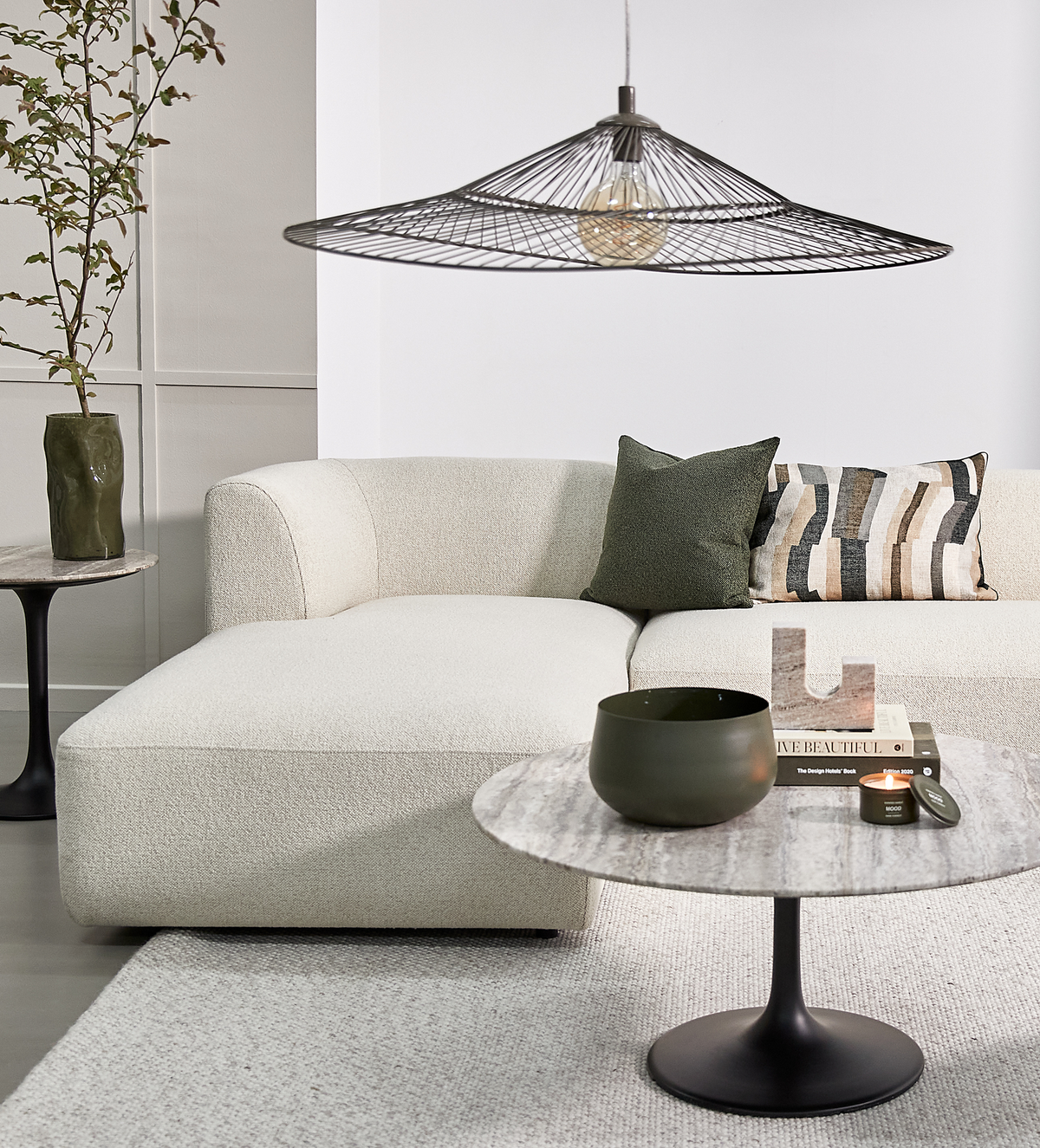 Lyst stue med cremefarvet sofa, trådpendel over og rundt marmorsofabord med grøn skål, tændt duftlys og bogen ”Live Beautiful”; grøn og mønstret pude samt vase med grene.