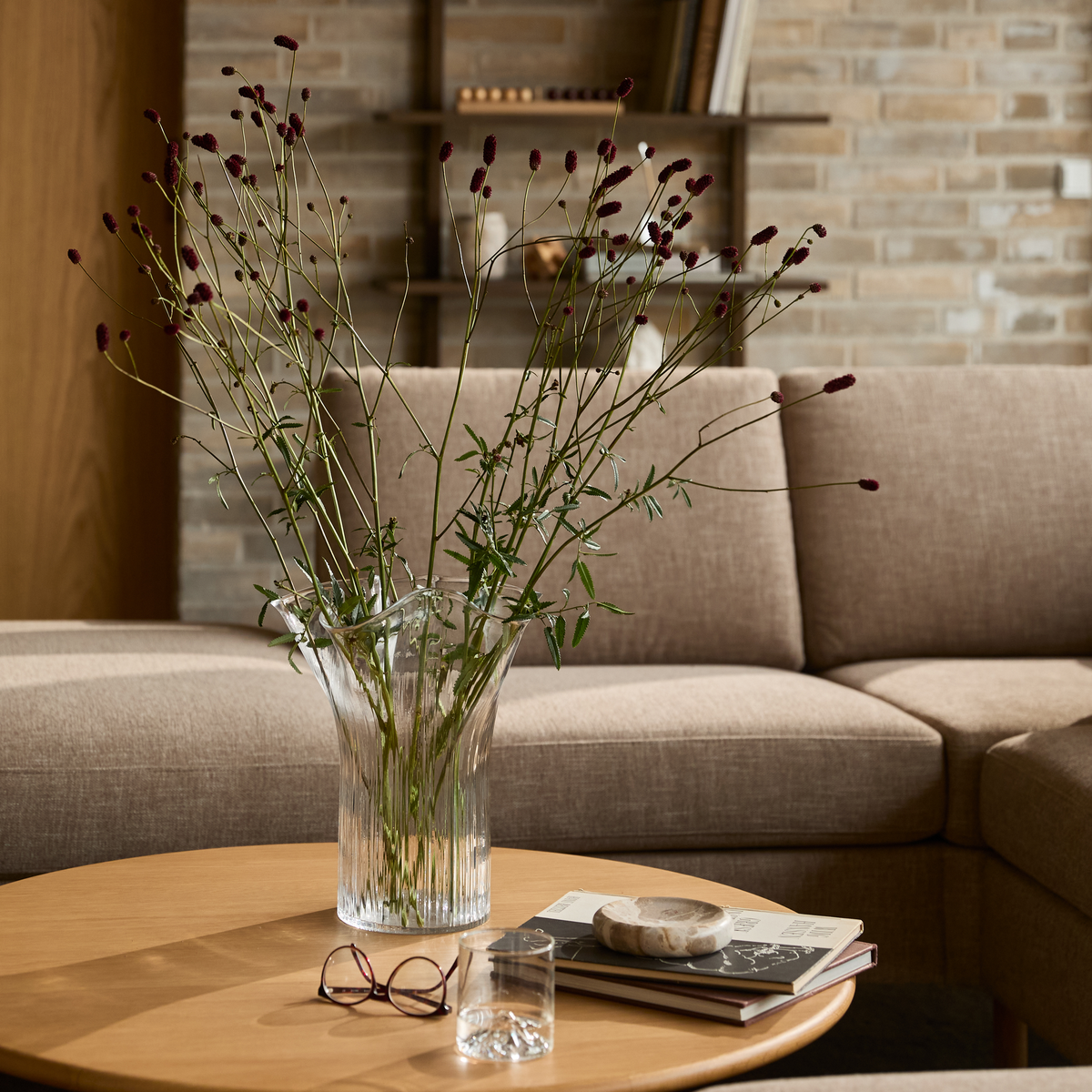 Glasvase med lange grene på et rundt træbord foran en beige sofa; bøger, et drikkeglas og briller ligger på bordet.