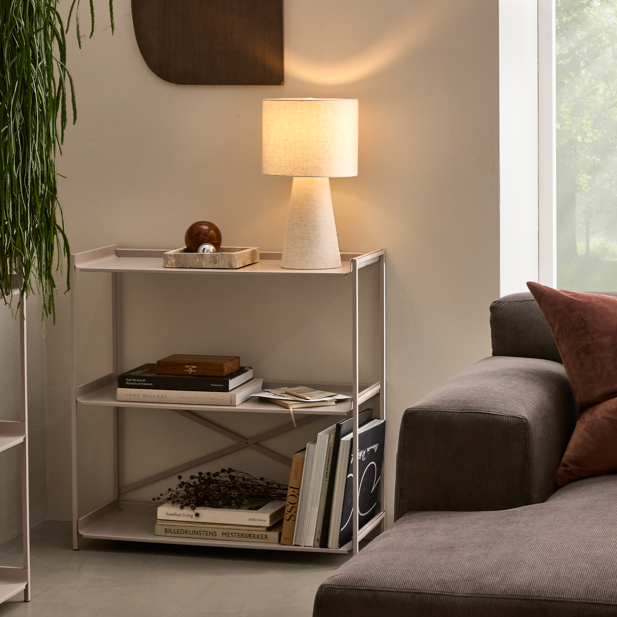 Lyst og neutralt stuehjørne med grå sofa og brune puder, lille reol med tænd bordlampe og bøger; en synlig titel er “BILLEDKUNSTENS MESTERVÆRKER”.