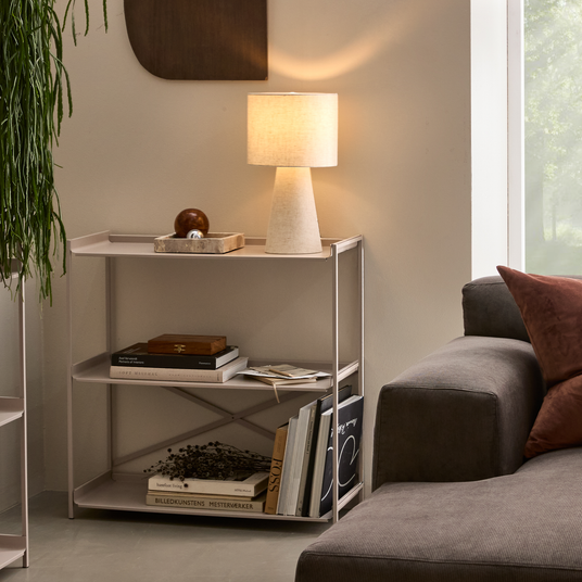 Lyst og neutralt stuehjørne med grå sofa og brune puder, lille reol med tænd bordlampe og bøger; en synlig titel er “BILLEDKUNSTENS MESTERVÆRKER”.