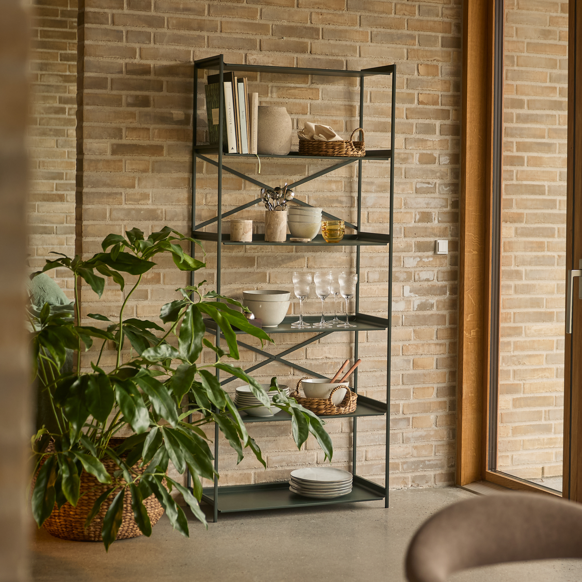 Metalthylde mod murstensvæg med keramik, glas og kurve, stor grøn potteplante ved siden af i dagslys.