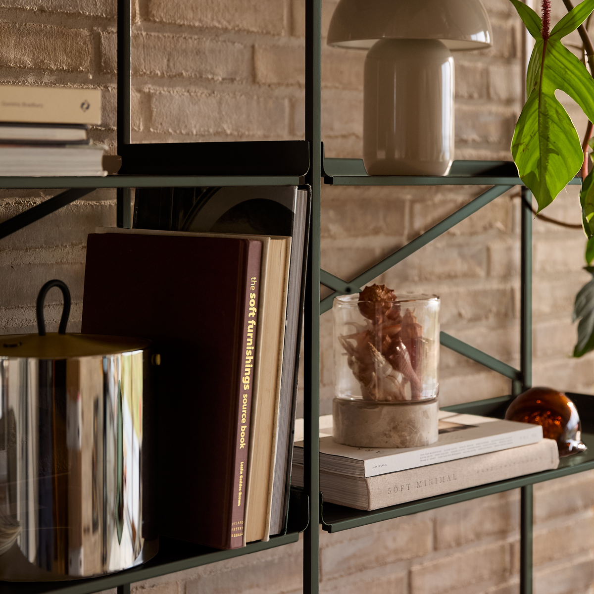 Stylet metalreol mod murstensvæg med bordlampe, grøn plante og bøger som ”the soft furnishings source book” og ”SOFT MINIMAL”, samt glasbeholder med tørrede planter.