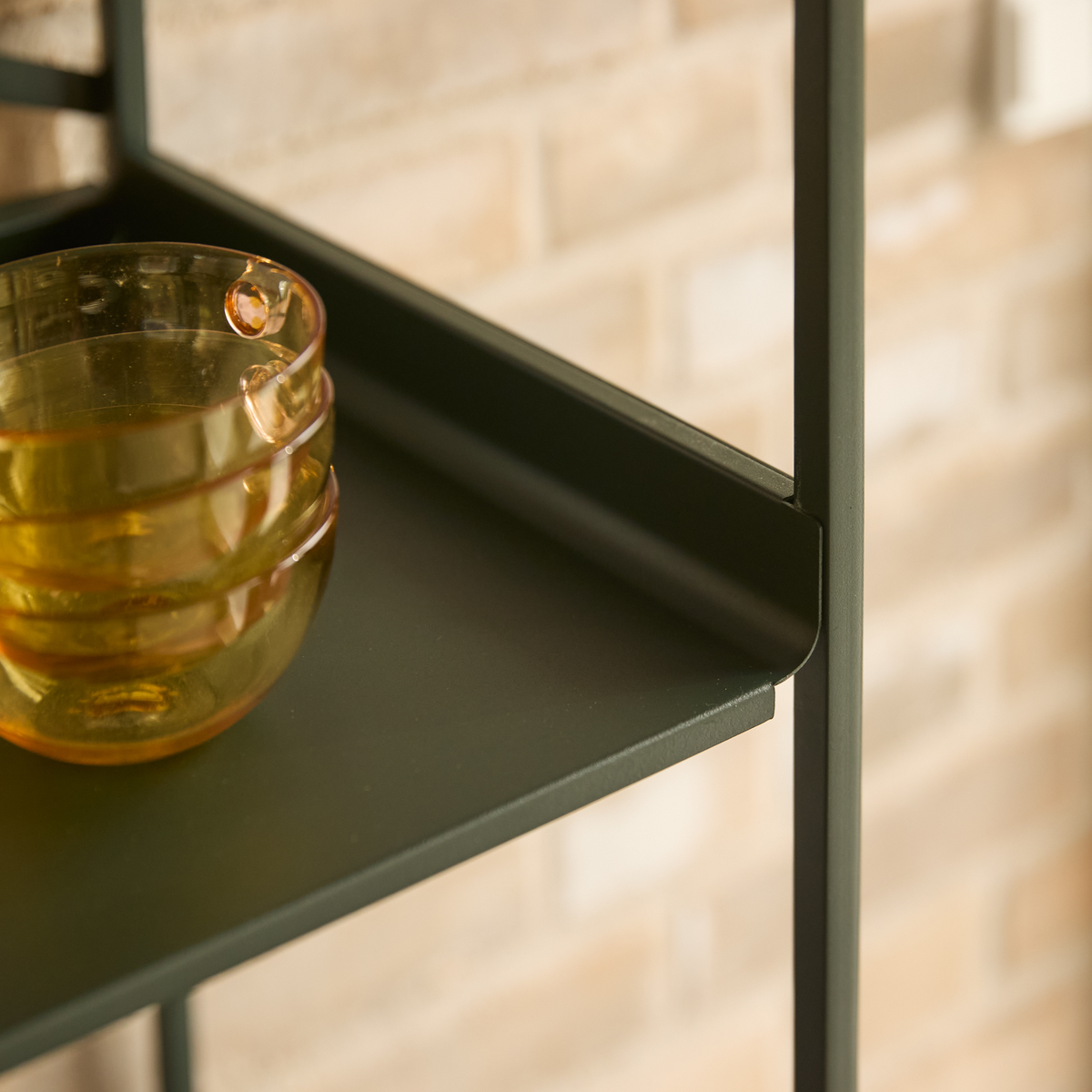 Nærbillede af olivengrøn metalhylde med stablede ravfarvede glasskåle mod beige murstensvæg.