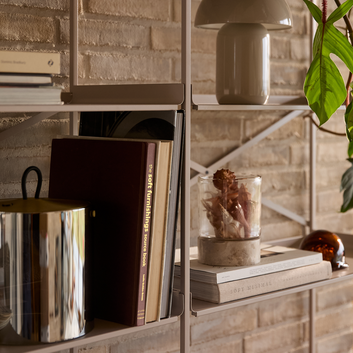 Nærbillede af en stylet reolsektion med bøger, bl.a. ”the soft furnishings source book” og ”SOFT MINIMAL”, en lille keramisk lampe, et glas med tørrede planter, en blank metalbeholder og en grøn plante mod en murstensvæg.